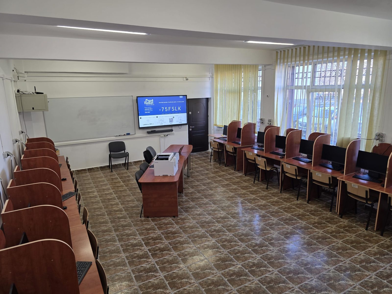 Laboratorul de informatică dotat prin proiect: șiruri de mese cu stații de lucru, tablă interactivă pe peretele din față